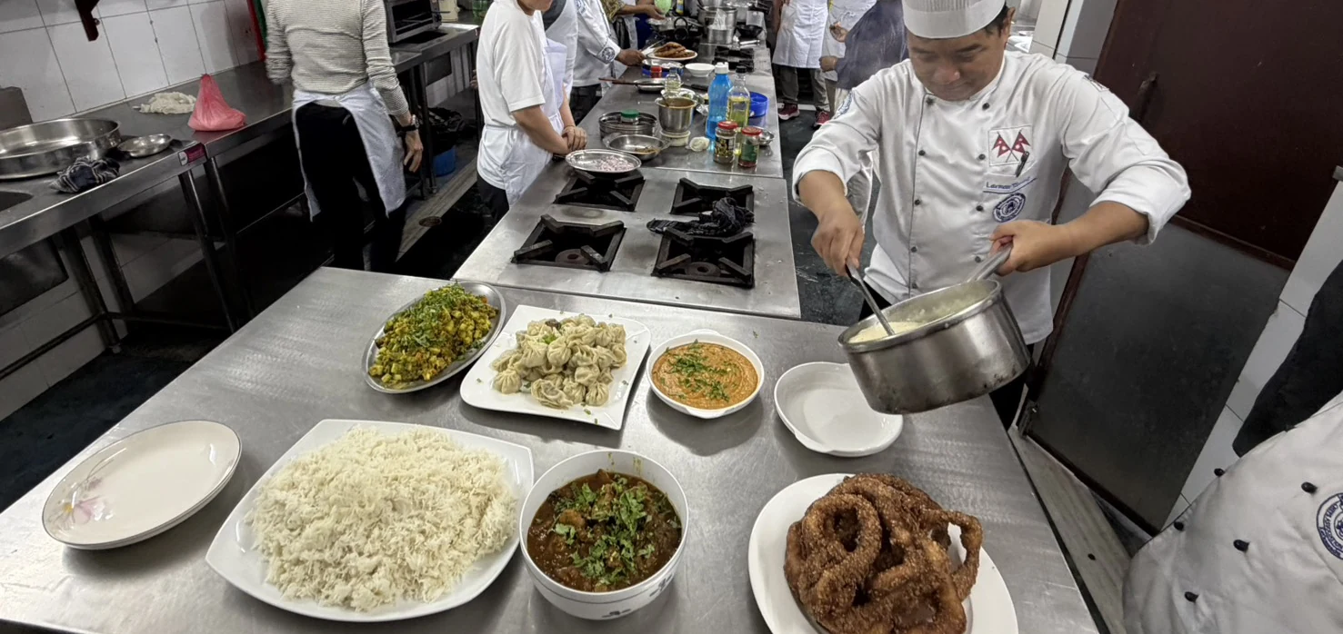 料理学校の調理台に並んだダルバート、モモ、カレー、セルロティ。奥で料理人が鍋から盛り付けている